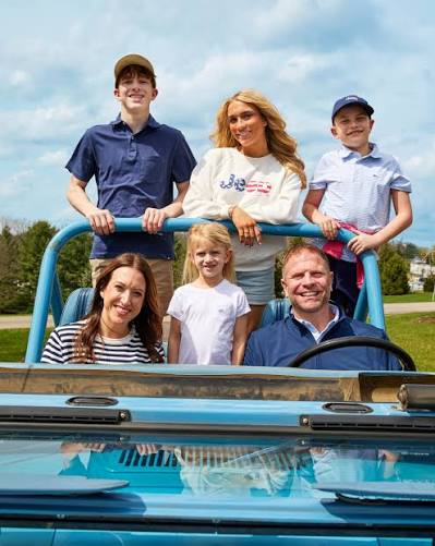 Family of six smiling in a vintage blue convertible on a sunny day